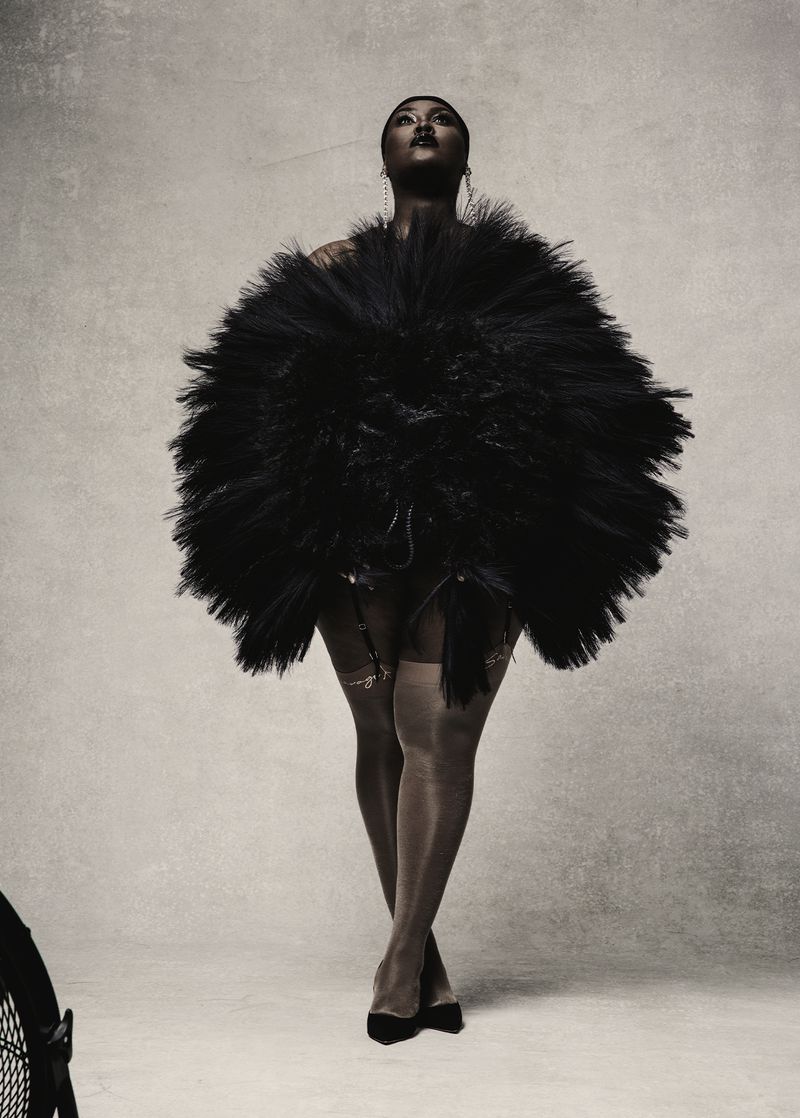 Full shot of a black woman posing in a sculptural black dress with hose and heels in a photo studio with soft lighting and a neutral backdrop.