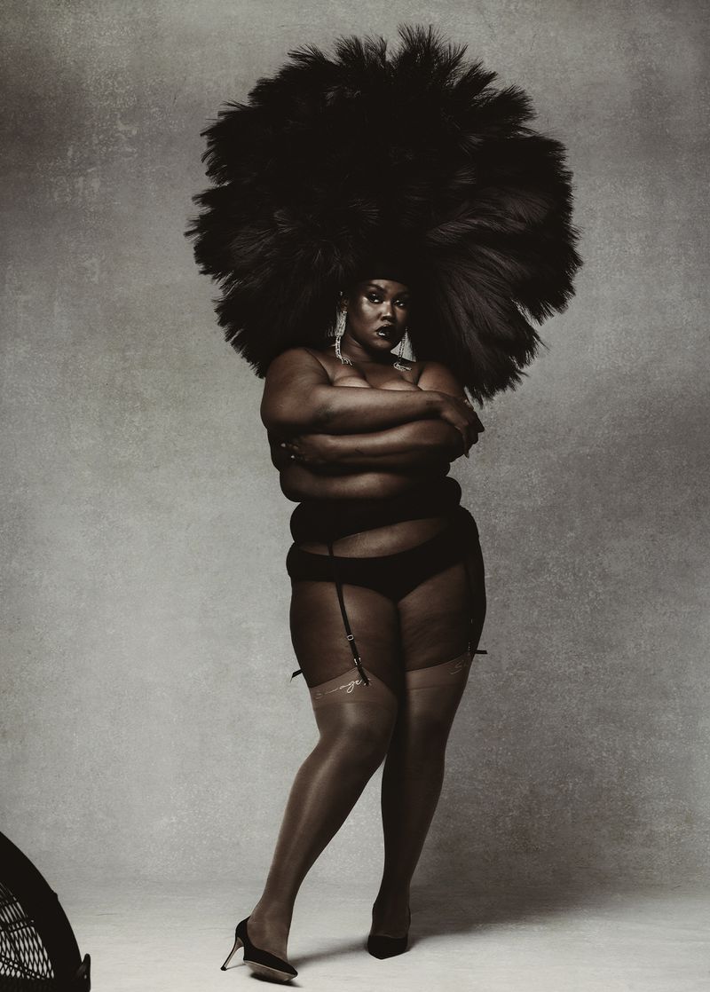 Full-length studio shot of a black woman in lingerie and heels, wearing a voluminous afro hairstyle, arms crossed, and expressionless.