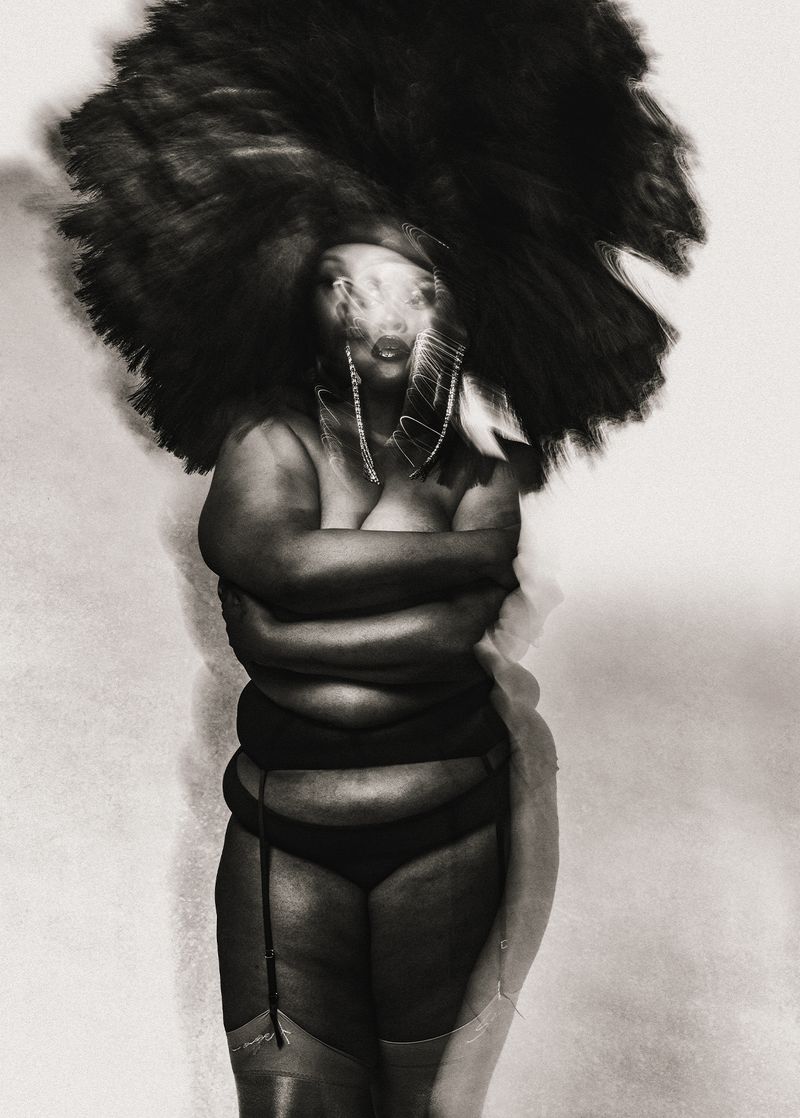 Portrait of a plus-size African-American woman with arms crossed, wearing lingerie and a large, dark afro wig against a plain backdrop.