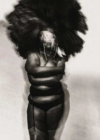Portrait of a plus-size African-American woman with arms crossed, wearing lingerie and a large, dark afro wig against a plain backdrop.