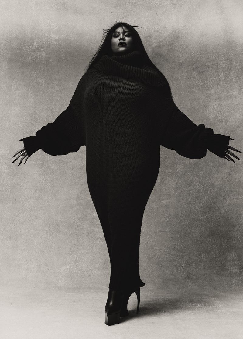 A Black female fashion model poses in a dark turtleneck sweater dress with long sleeves against a neutral backdrop in a studio shot.