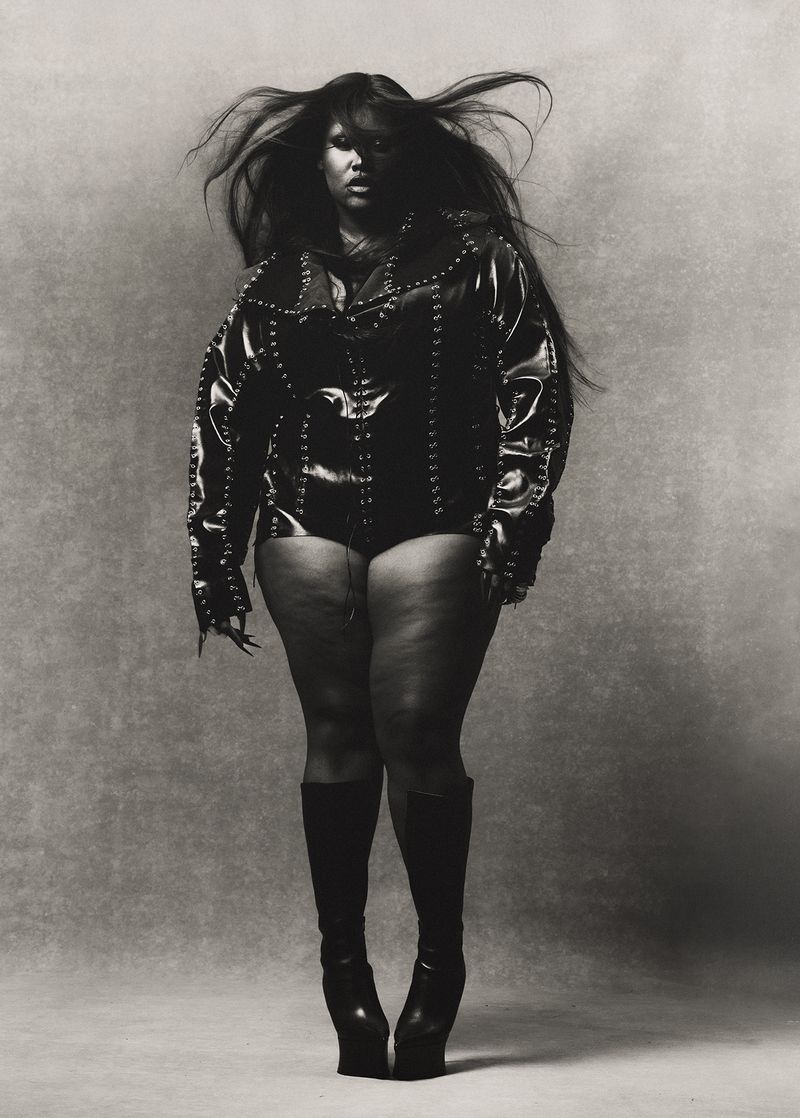 Monochrome portrait of Lizzo wearing a studded jacket, bodysuit, and high platform boots, posing in front of a neutral backdrop.