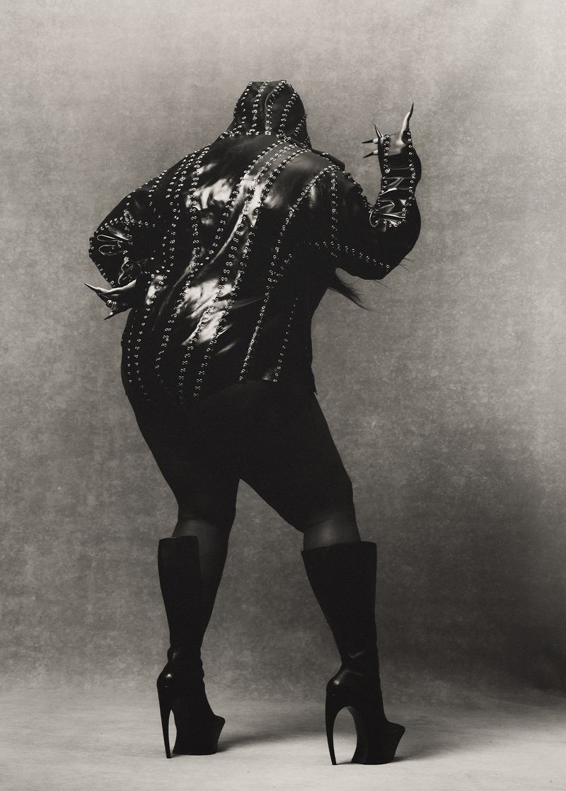 A person stands with back to the camera in a studded jacket, skirt, and heeled boots, holding up their hand in front of a grey backdrop.