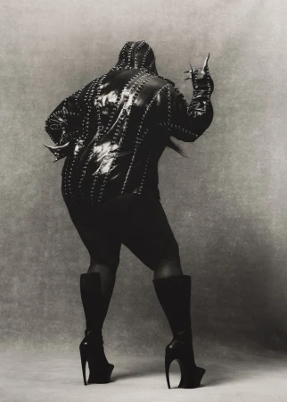 A person stands with back to the camera in a studded jacket, skirt, and heeled boots, holding up their hand in front of a grey backdrop.