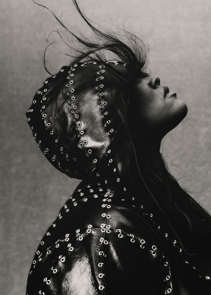 Monochromatic fashion portrait of a person looking up wearing a studded leather head covering and jacket against a grey background.