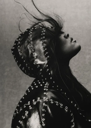 Monochromatic fashion portrait of a person looking up wearing a studded leather head covering and jacket against a grey background.
