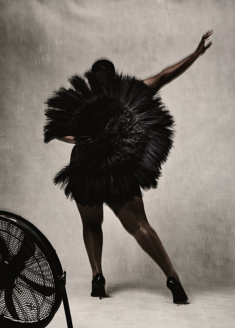 Woman in black feathered dress poses in a studio with a retro fan. Studio lighting, high heels, and neutral backdrop create a striking image.