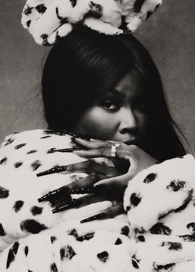 Black and white portrait of a woman with long black nails, wearing a dalmatian print coat and bow, gazing at the camera.