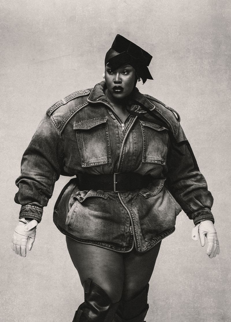 Stylish plus-size woman in denim jacket and black cap confidently poses against a simple light background in a black and white fashion shot.
