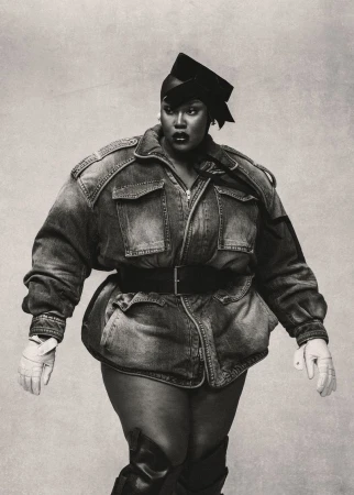 Stylish plus-size woman in denim jacket and black cap confidently poses against a simple light background in a black and white fashion shot.