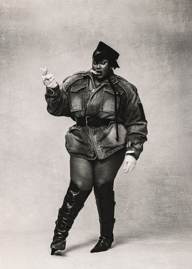 A portrait captures a plus-size African-American woman wearing a denim outfit and black leather boots, smoking against a neutral background.