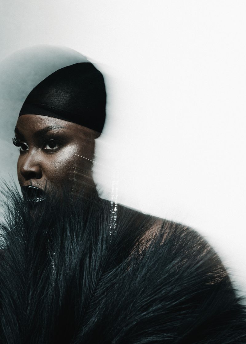 Close-up portrait of a Black woman with dramatic makeup and a black headscarf, wearing a feathered garment against a light backdrop.