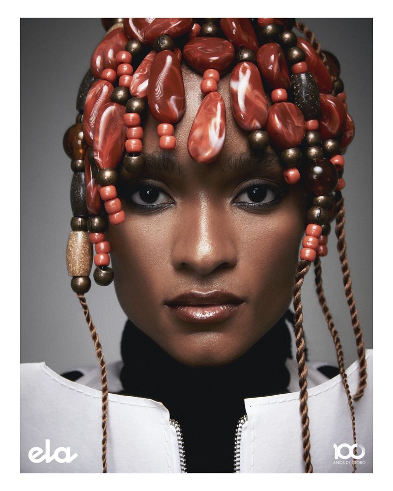 A headshot of a woman with a bead headdress, wearing a white jacket over a black turtleneck, against a gray background.