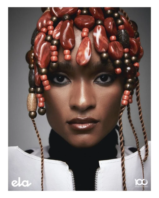 A headshot of a woman with a bead headdress, wearing a white jacket over a black turtleneck, against a gray background.
