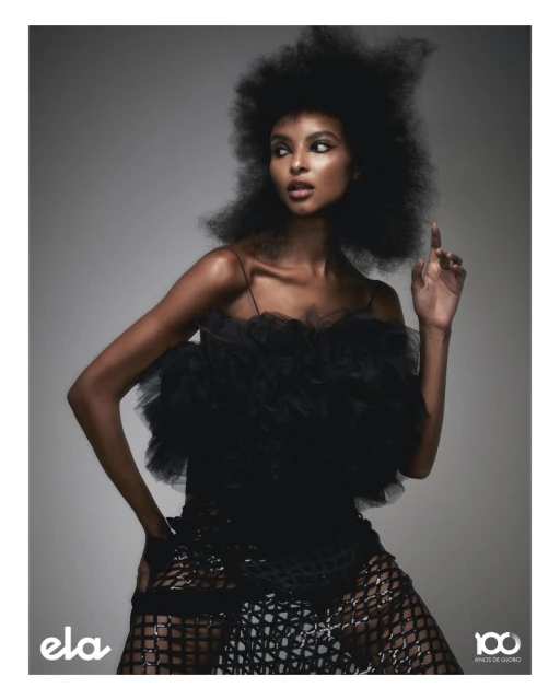 Woman with afro hairstyle poses in a black dress with a ruffled bodice in front of a studio backdrop, fashion portrait.