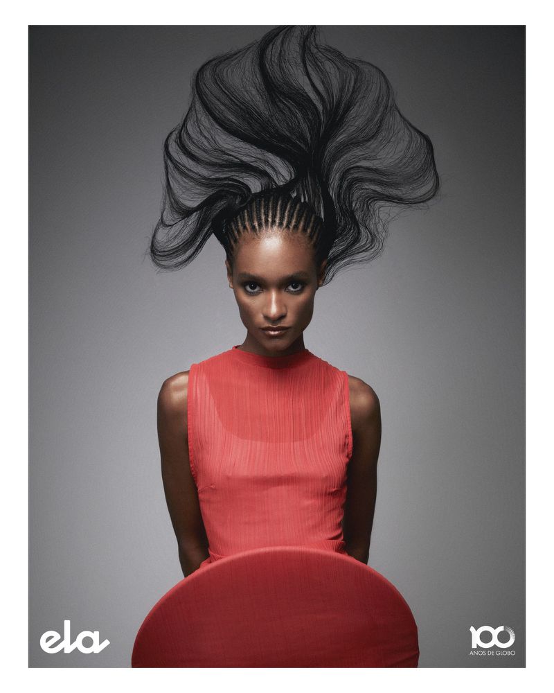 Headshot of an African American woman with elaborate braided hairstyle, wearing a coral dress, posing in front of a gray background.