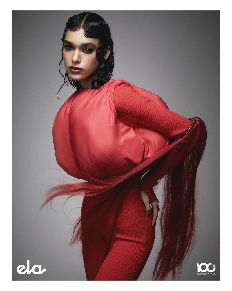 Fashion portrait of a woman with dark hair in a sculptural red dress against a grey studio backdrop in a vertical composition.