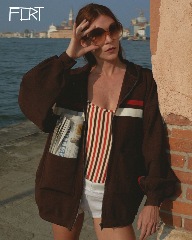 A woman with sunglasses and a striped top stands by a canal in a brown jacket with a newspaper in the pocket, city buildings in the background.