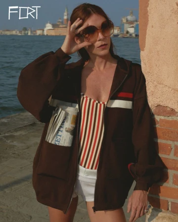 A woman with sunglasses and a striped top stands by a canal in a brown jacket with a newspaper in the pocket, city buildings in the background.