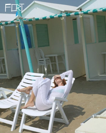 A woman rests on a white beach chair near white beach cabanas under the daytime sun, enjoying a relaxing day at the beach.