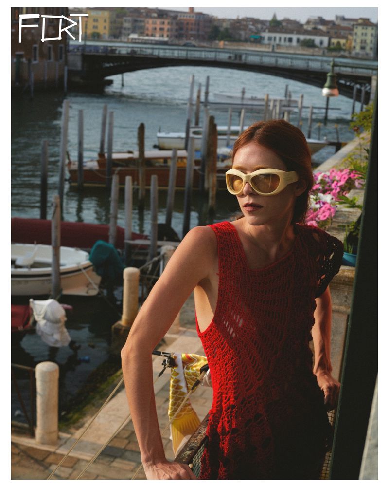 Woman in a red dress and cream sunglasses poses on a balcony overlooking a canal with boats and buildings in Venice, Italy.