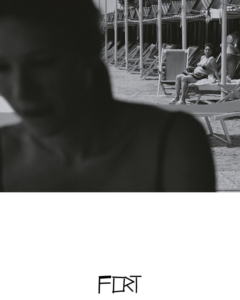 Black and white photo of a woman sitting on a beach chair, with other chairs and shelters in the background and another person in foreground.