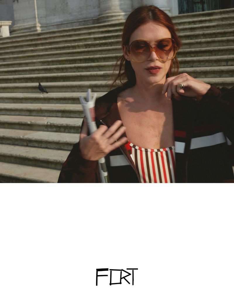 Adult woman in a brown jacket poses in front of large stairs with a newspaper in hand; a pigeon is visible on the steps.