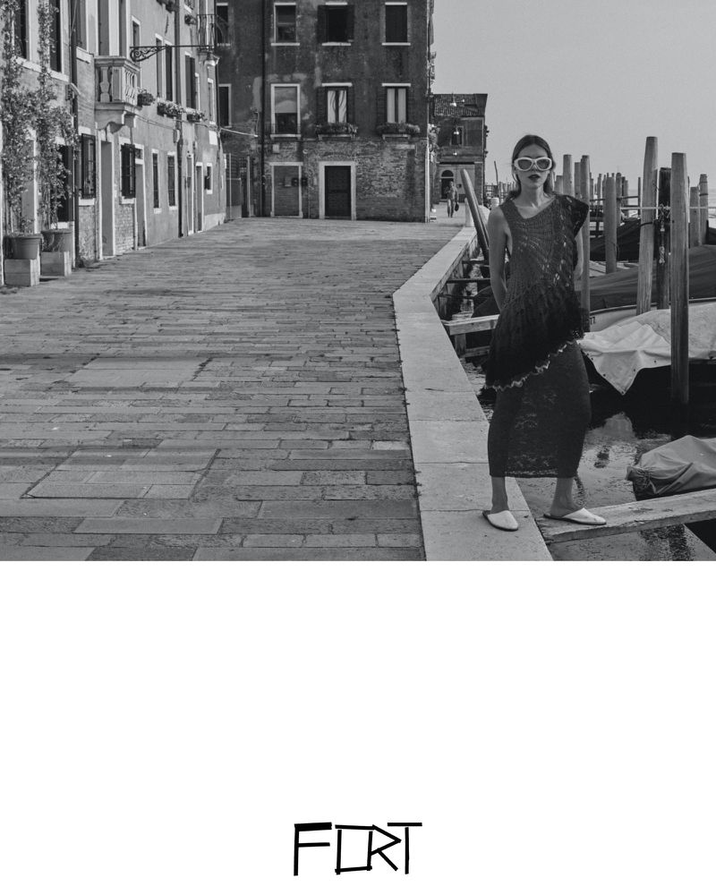 Black-and-white fashion shot of a woman with sunglasses in a sleeveless outfit, posing on a Venice canal-side walkway with buildings.
