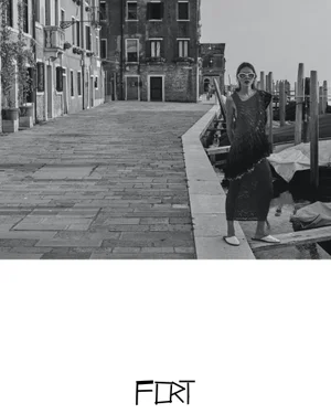 A woman wearing sunglasses and a dark knit dress stands along a stone canal pathway in Venice in black and white.