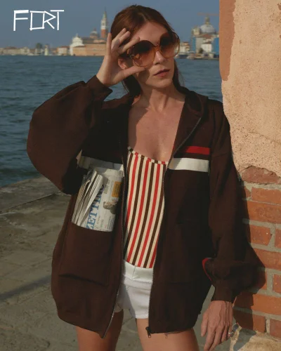 A woman in a brown retro jacket, striped top, and oversized sunglasses leaning against a brick wall by a Venice waterfront.