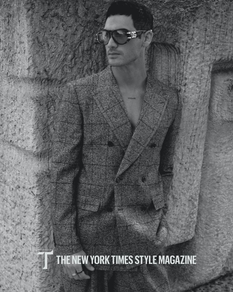 Monochrome portrait of a stylish adult male model in a checkered suit and sunglasses, posing against a stone wall for a magazine.