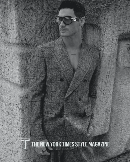 Monochrome portrait of a stylish adult male model in a checkered suit and sunglasses, posing against a stone wall for a magazine.