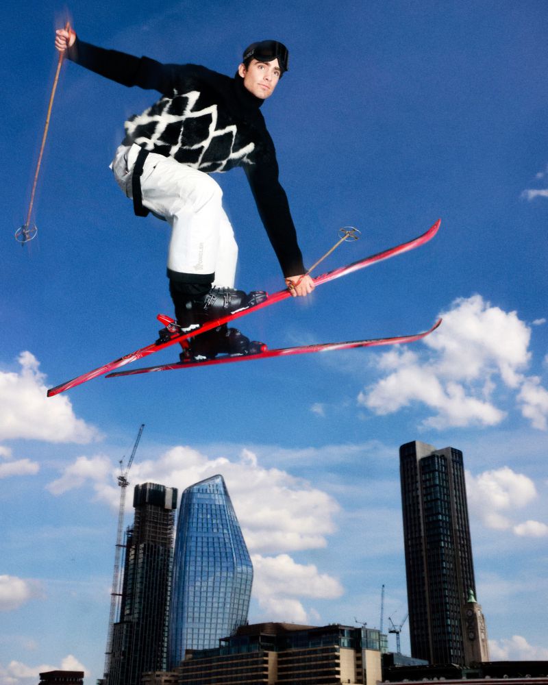 A man is jumping in mid-air on skis over buildings. He is wearing a black and white sweater and white pants. Blue sky in the background.