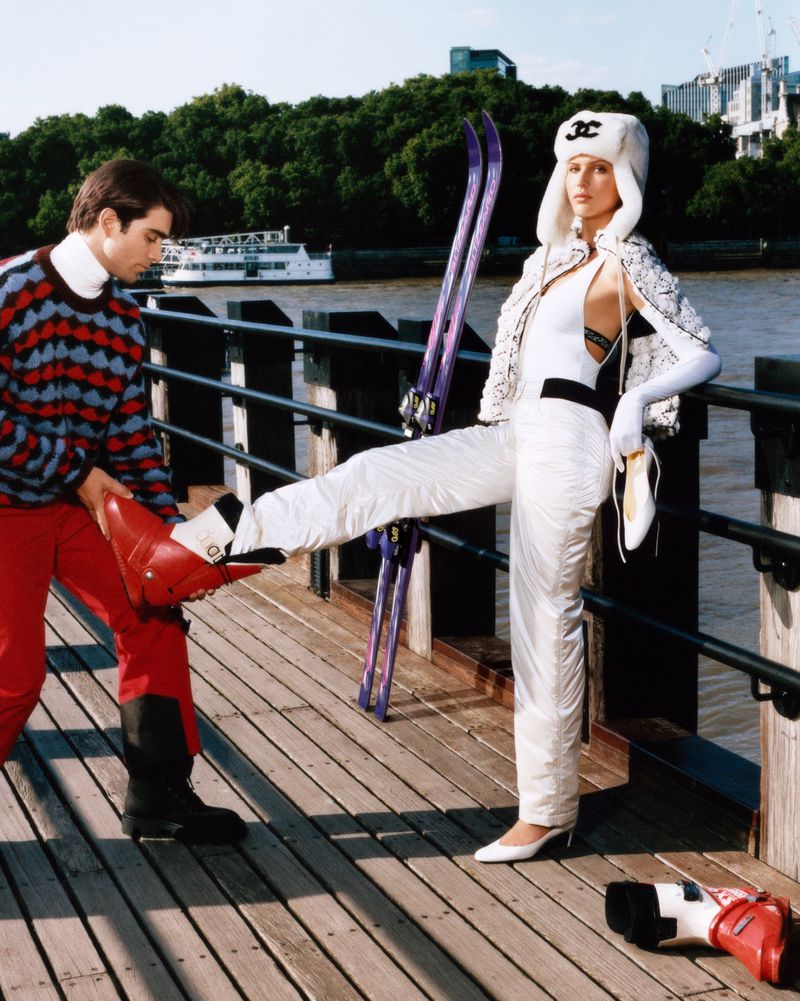 Fashion shot of two people, one adult female in ski outfit holding skis, and one adult male assisting with putting on ski boots on wooden dock.