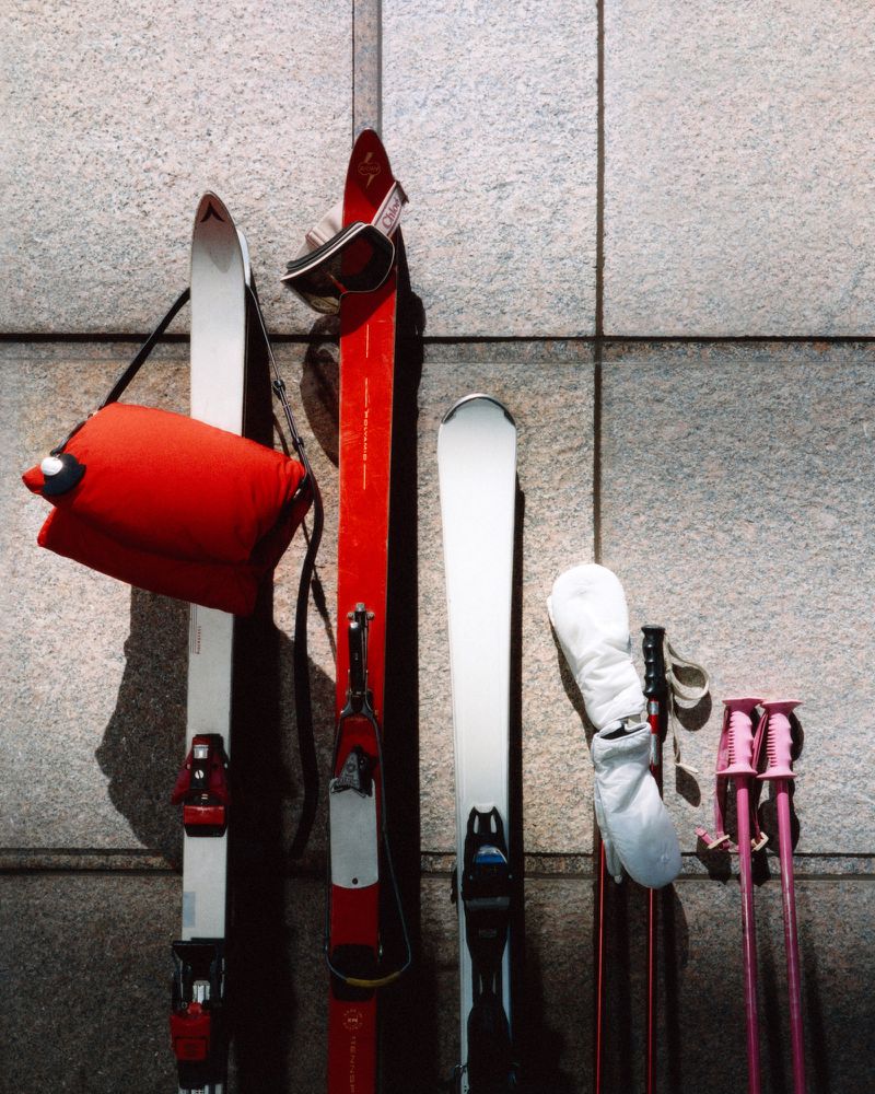 Skis, ski poles, goggles, and a red bag stand against a textured grey wall, a still life arrangement of winter sports equipment.