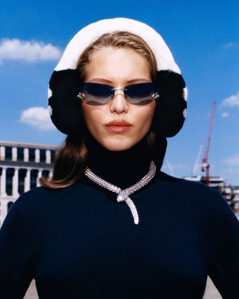 Portrait of a young woman wearing earmuffs, sunglasses, and an elaborate Bulgari Serpenti necklace, with urban buildings in the background.