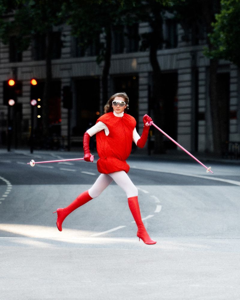 A woman in a red outfit runs across a city street with ski poles, creating a high-fashion, whimsical urban image.