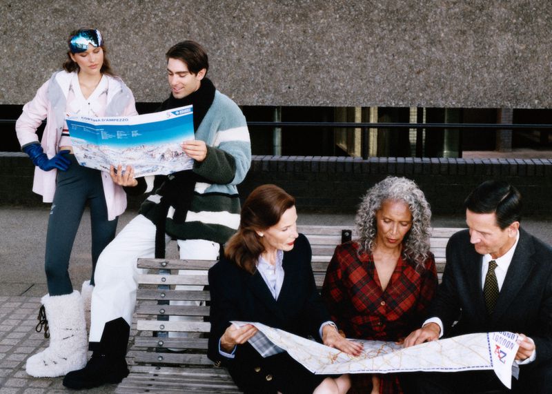 Group of people gathered outdoors on bench with winter clothing and maps, city environment with textured concrete wall in the background.