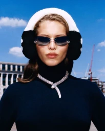 Model wearing dark sunglasses, a black turtleneck, snake necklace, and white earmuffs against a blue sky.