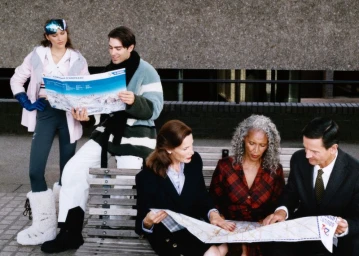 Models in winter skiwear and tailored suits reading travel maps while gathered around a bench against a textured concrete wall.