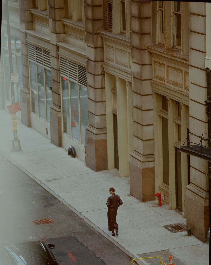 A woman in dark clothing walks on a sidewalk next to a city street and buildings, captured from a high-angle, eye-level perspective.