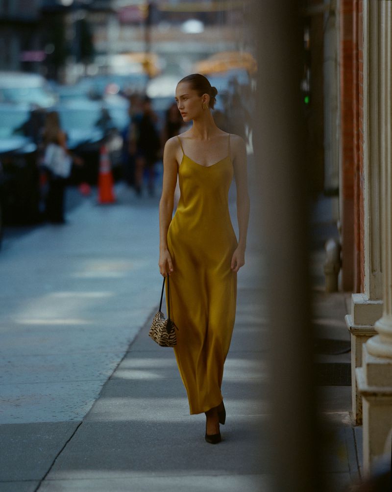 Woman in yellow silk dress walking on city sidewalk with leopard-print purse. Stylish urban fashion scene. Pedestrians in background.