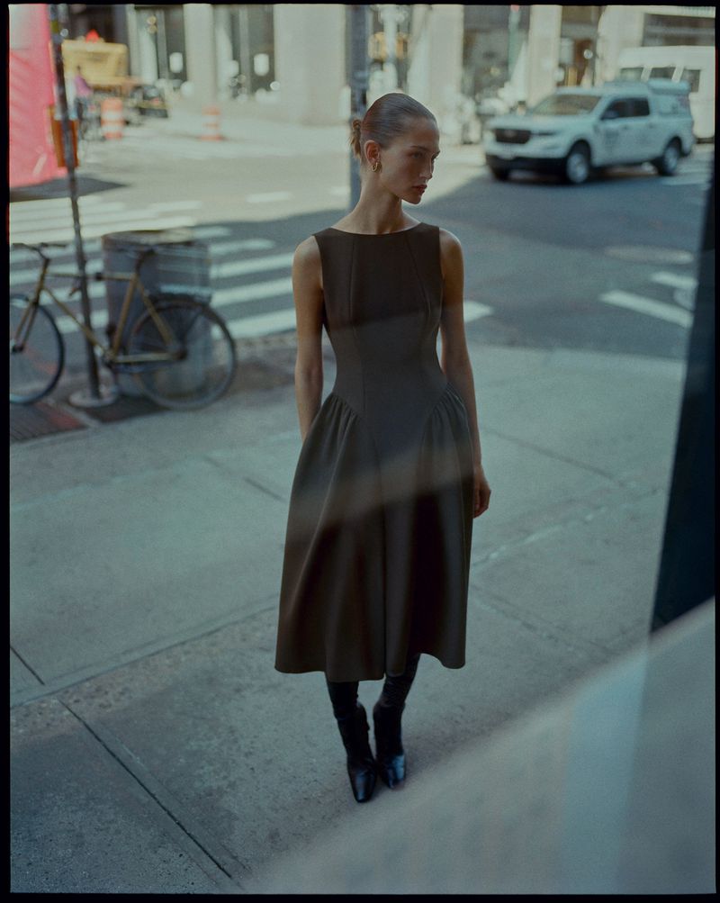 Woman in a brown dress and black boots standing on a city street with a bicycle and white SUV in the background, reflecting on glass.
