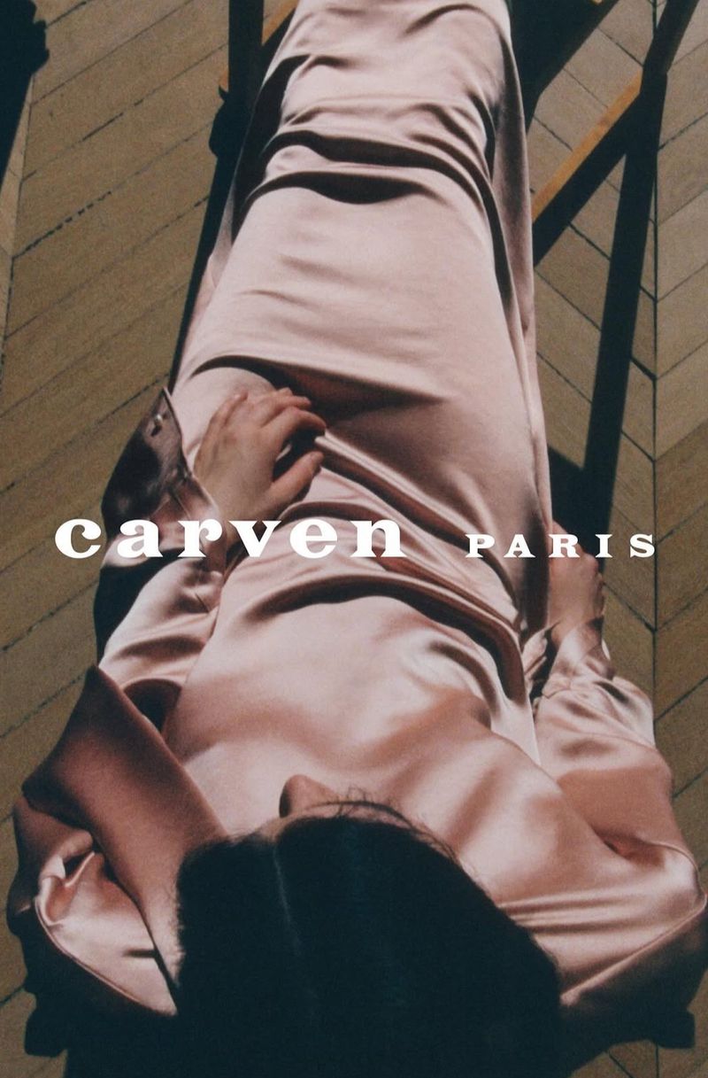 Top-down fashion shot: a person in a pink satin dress relaxes on a wooden chair, sunlight highlighting the fabric. 'CARVEN PARIS' text overlay.