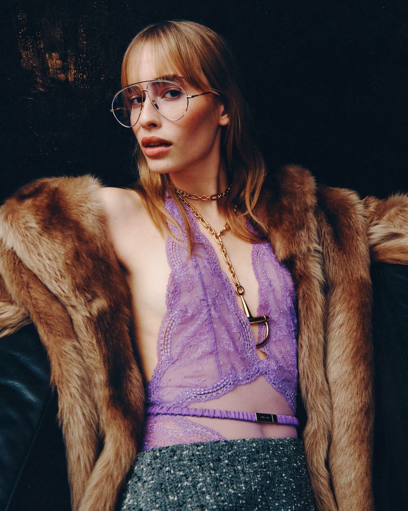 A woman in purple lingerie and glasses poses with a fur coat, set against a dark background in a studio shot.