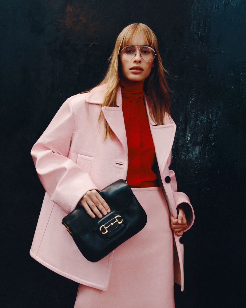 A poised woman models a pink skirt suit with a black purse against a textured dark background, emphasizing fashion and style.