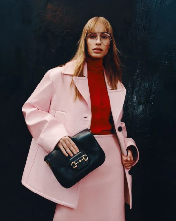 A poised woman models a pink skirt suit with a black purse against a textured dark background, emphasizing fashion and style.
