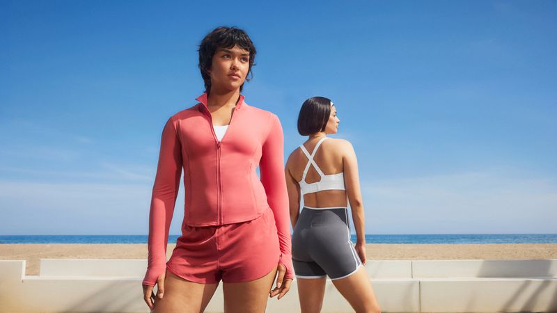 Two women wearing athletic sportswear standing near a beach under a clear blue sky.