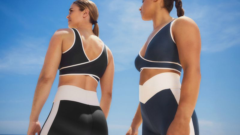 Two women in athletic wear pose outdoors against a bright blue sky, showcasing active lifestyle and fitness apparel, with focus on body positivity.