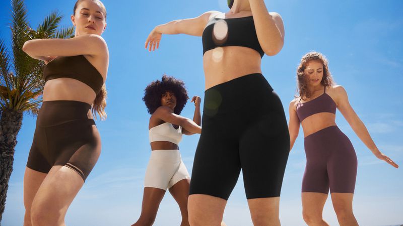 Four diverse women in athletic wear pose outdoors against a blue sky with a palm tree in the background, showcasing fitness apparel.